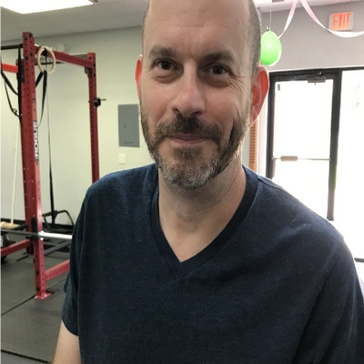 Man with a beard in a gym setting with workout equipment behind him.