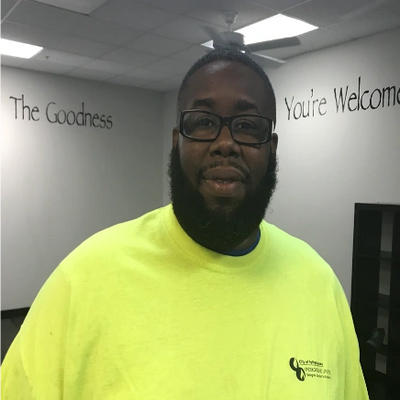 Man wearing glasses and a bright yellow shirt stands indoors with a welcoming message on the wall.