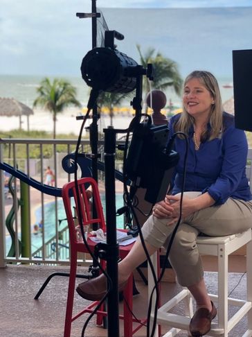 Woman in blue shirt sitting on a white chair with filming equipment around her at a beachside location.
