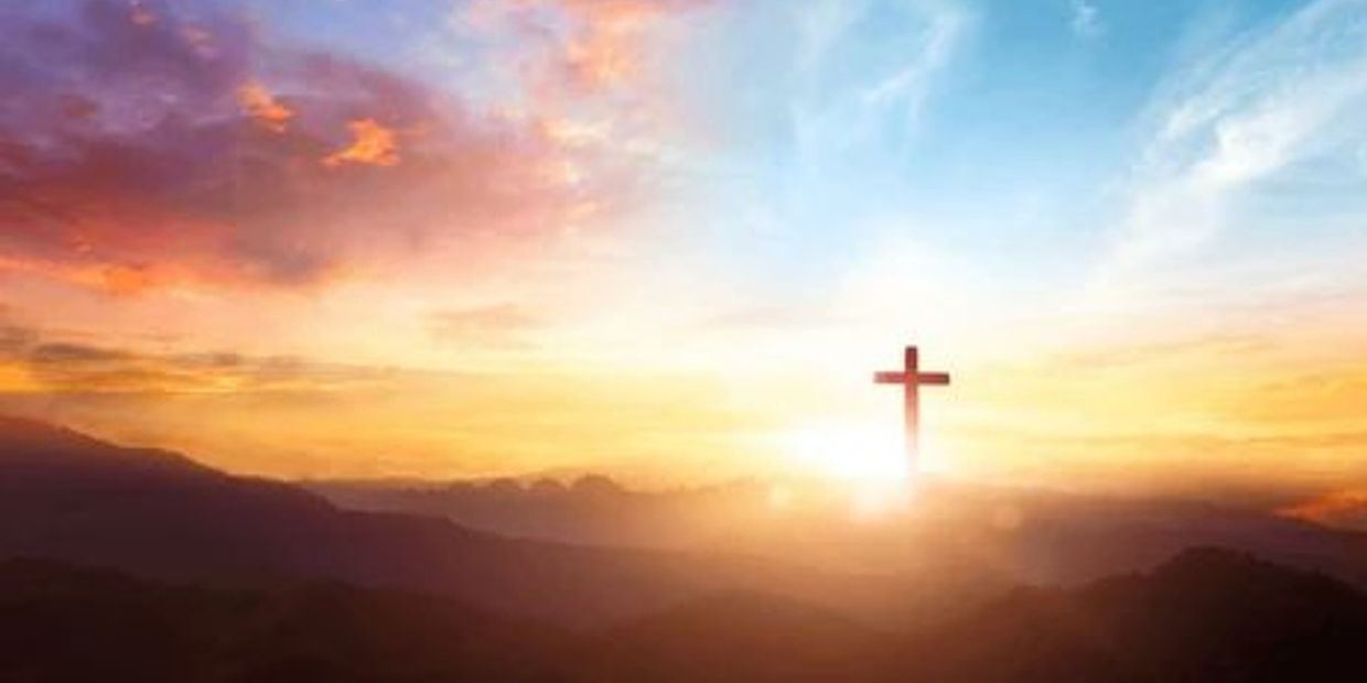 A glowing cross illuminated by a colorful sunrise over misty mountains.
