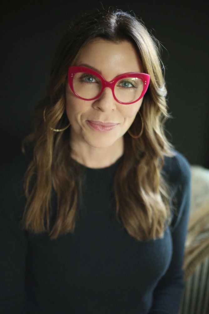 Woman with long brown hair wearing bold red glasses and a black top smiles gently.