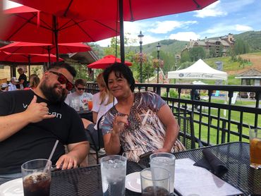 Two people smiling and making a shaka sign at an outdoor restaurant with red umbrellas.
