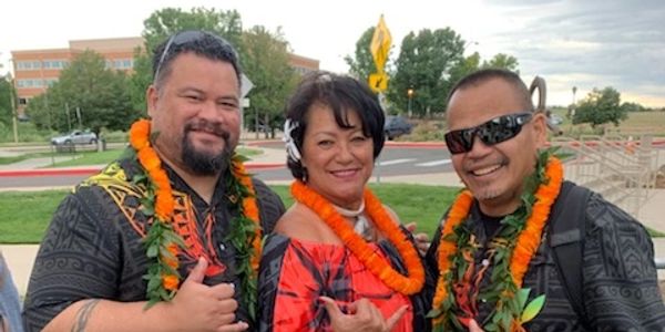 Three people wearing colorful Hawaiian shirts and leis posing outdoors with shaka signs.