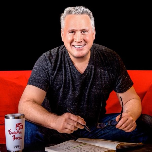 Man smiling while holding glasses and pen, sitting by a notebook.