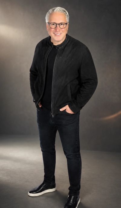 Smiling man in black jacket and glasses stands confidently.