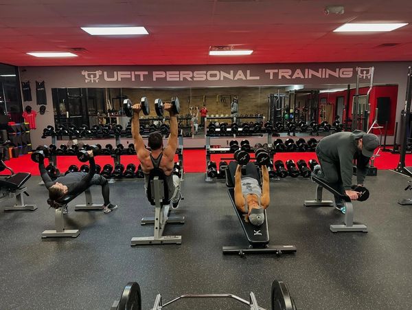Four people lifting dumbbells in a gym under "UFIT PERSONAL TRAINING" sign.
