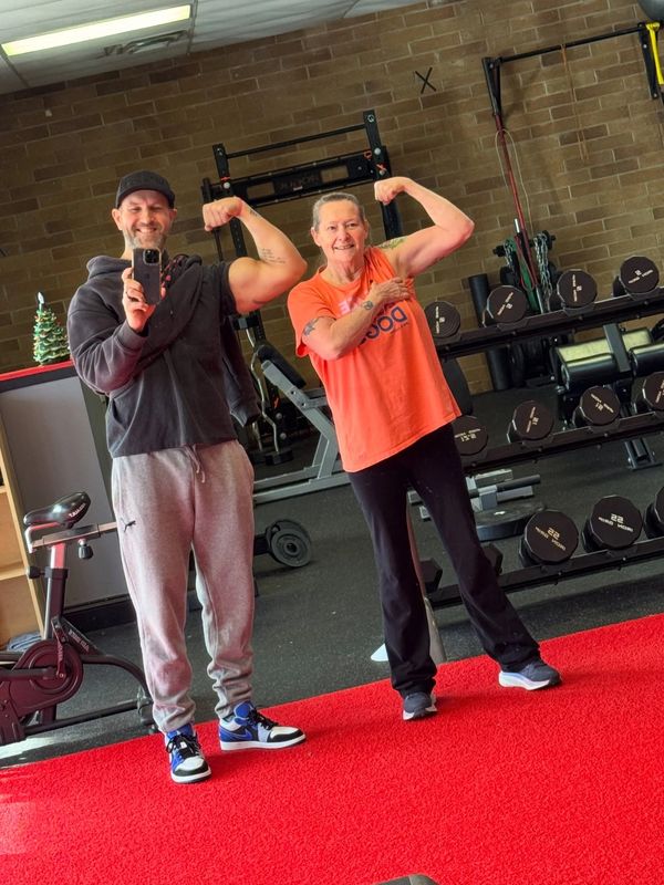 Two people flexing muscles in a gym mirror selfie.