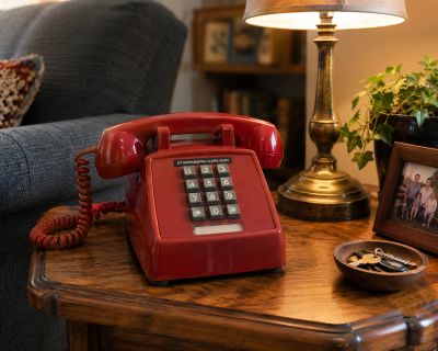 A vintage red rotary phone on a wooden side table with a lamp and framed photo.