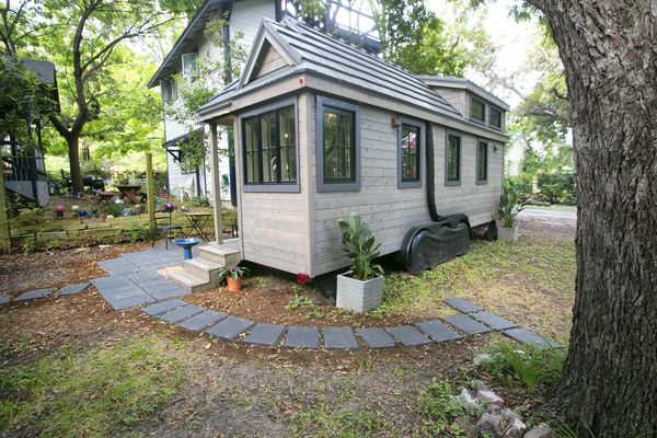 A compact tiny house with large windows nestled in a green, leafy yard.