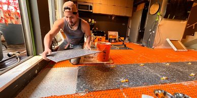 Man installing gray floor tiles on orange underlayment in a room under renovation.