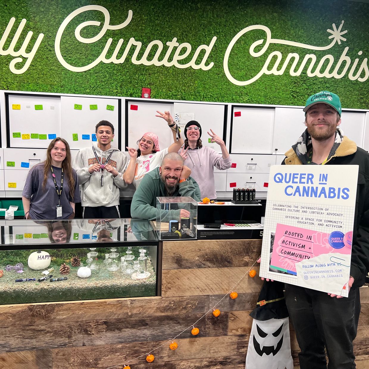 Group of smiling people in a cannabis store with a sign promoting queer advocacy in cannabis.