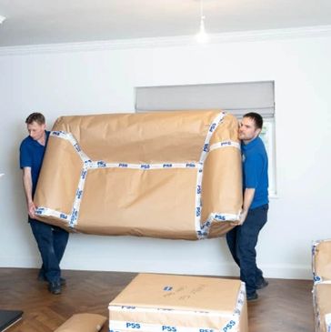 Two movers carrying a wrapped sofa inside a living room.