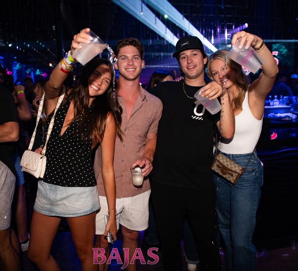 Four friends raising drinks and smiling at a lively party with colorful lights.
