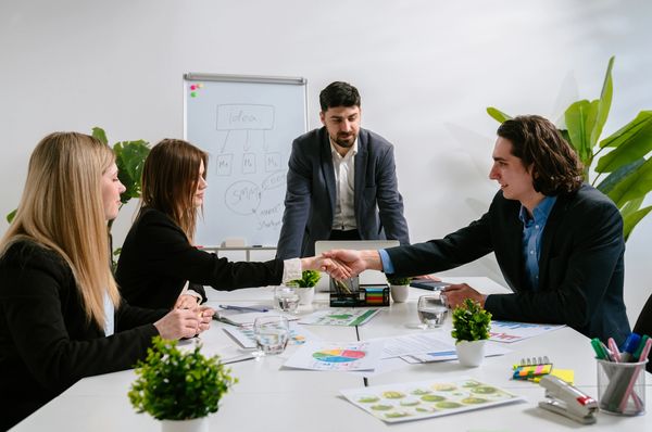 The image depicts a business meeting with four individuals around a white conference table.