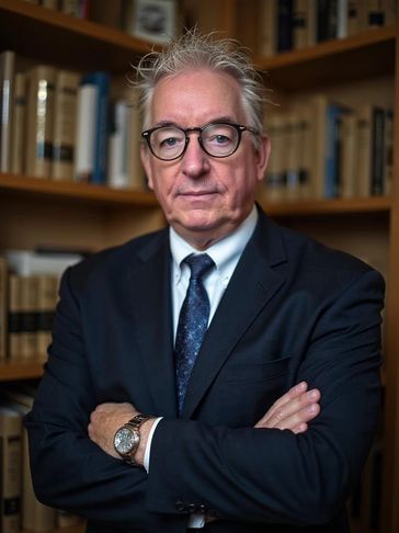 Elderly man in suit with glasses standing in front of bookshelves.