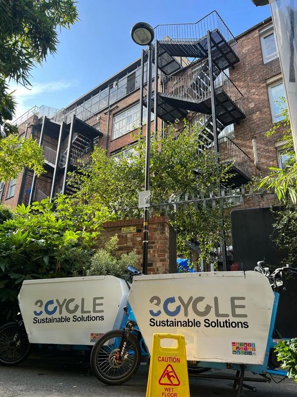Two cargo bikes with '3CYCLE Sustainable Solutions' parked near a brick building and fire escape.