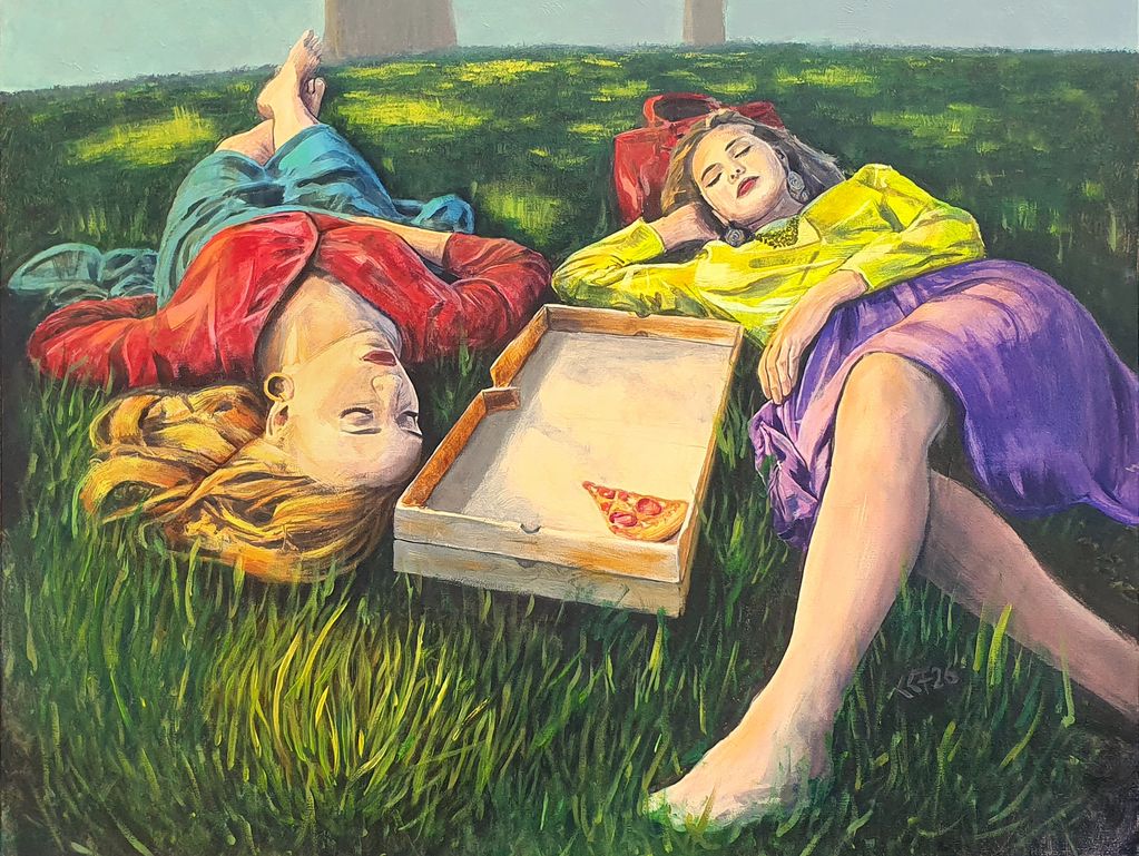 Two women lying on grass with a pizza box between them.