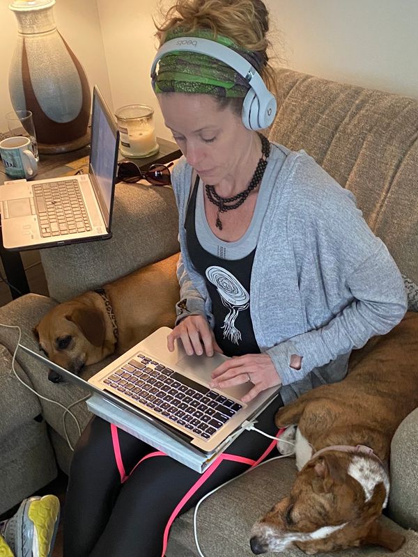Woman working on laptop with headphones, surrounded by two resting dogs on a couch.