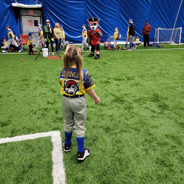 Young girl in baseball uniform watching a mascot on an indoor field.
