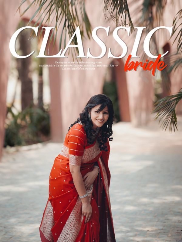 A smiling bride in a traditional red saree poses outdoors under palm leaves.