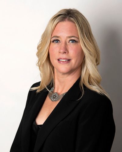 Professional portrait of a blonde woman in a black blazer with a statement necklace.