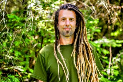 Man with long dreadlocks and green shirt stands in a lush forest.