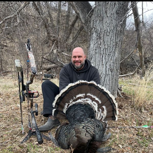 Hunter posing with a turkey and bow in a wooded area.