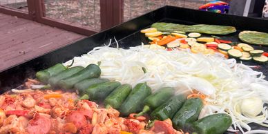 Grilled vegetables and marinated chicken cooking on a large outdoor griddle.