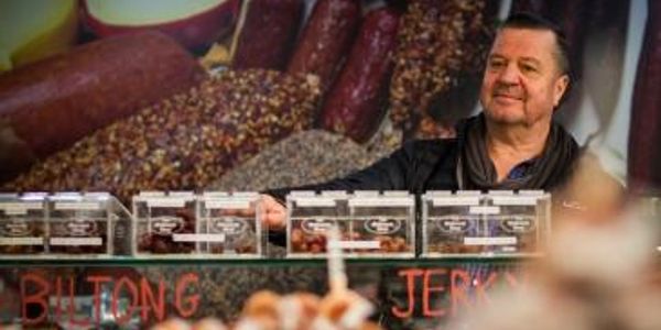 A vendor stands behind a display of packaged biltong and jerky at a market stall.