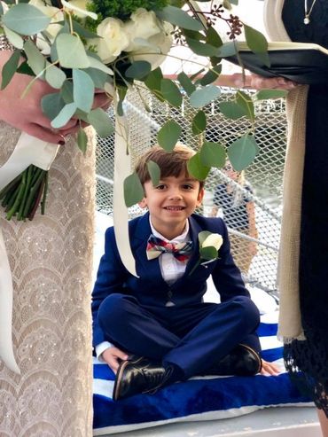 Vogm boy sitting at a wedding in a blue suit, colorful bow tie and white rose boutonniere.