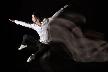 A male dancer leaps gracefully with motion blur against a dark background.
