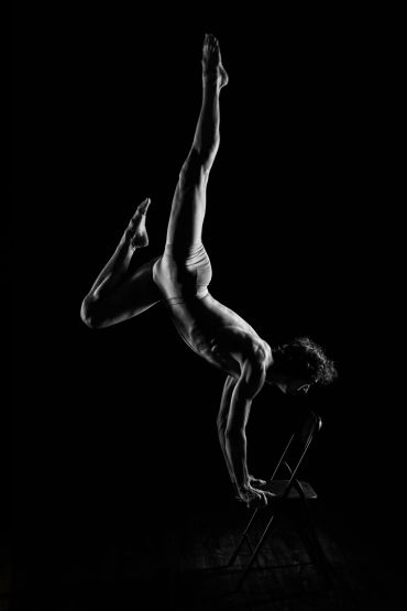 A dancer performs a handstand on a chair in dramatic lighting.