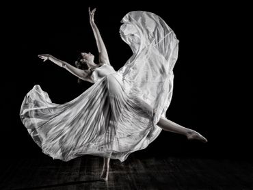Graceful dancer in flowing white dress leaps against black background.
