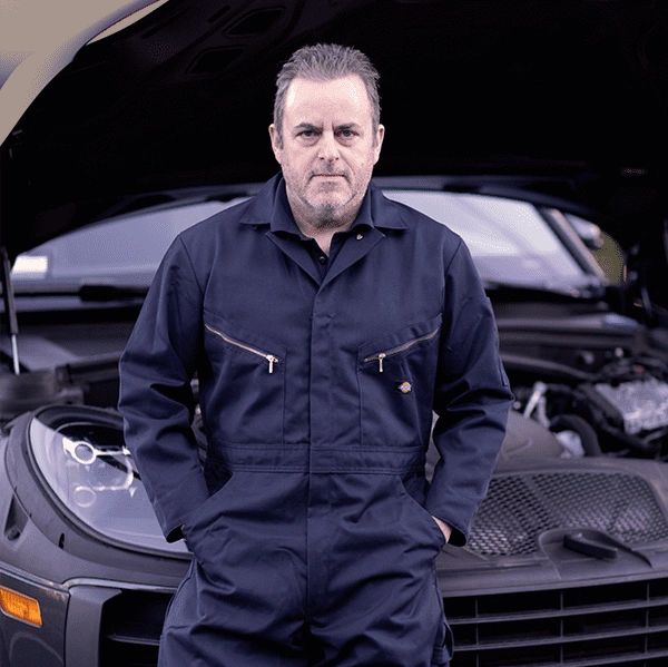 Mechanic in navy coveralls standing confidently in front of an open car hood.