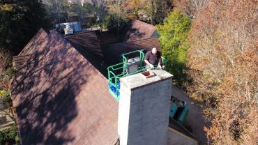 Worker inspecting chimney on a roof lift platform surrounded by autumn trees.