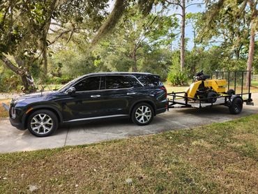 Black SUV towing a yellow machine on a trailer in a wooded driveway.