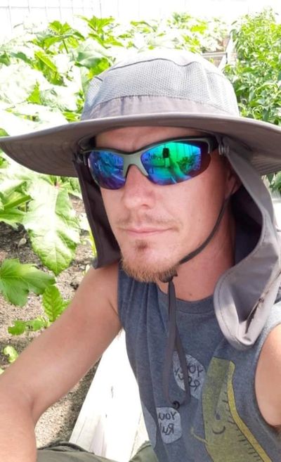 Photo of Justin, a light skinned man, wearing aviator sunglasses, a grey brimmed hat, and a tanktop.