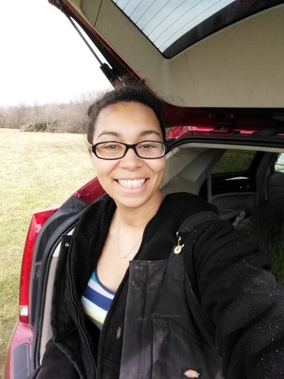 Photo of Jennifer, a medium skin toned person sitting on the edge of the trunk of a red car.