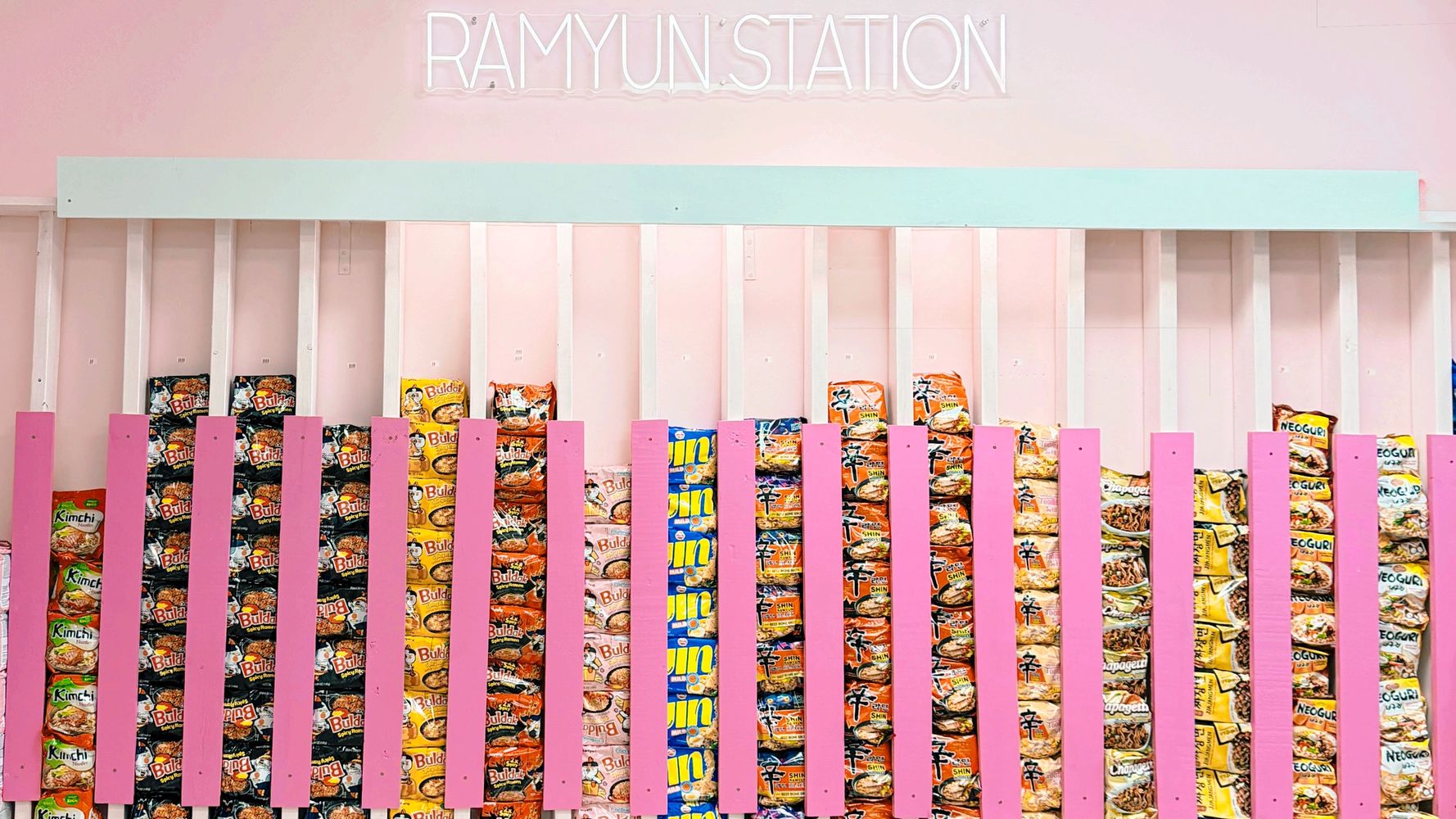 Colorful instant ramen packets displayed behind pink slats with a neon 'RAMYUN STATION' sign.