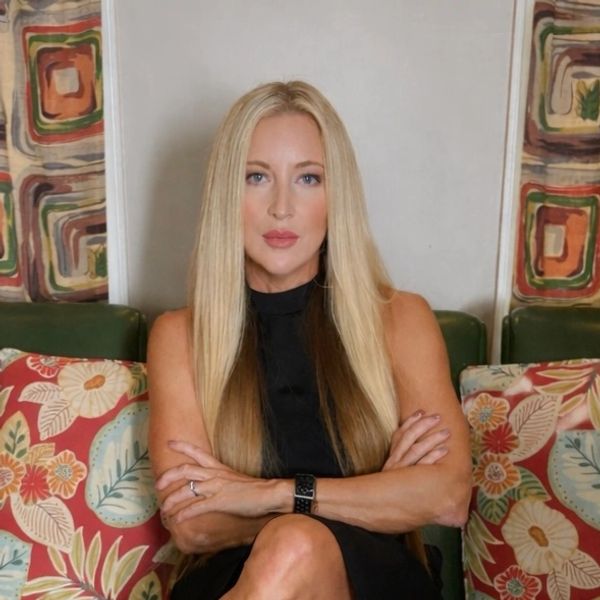 Blonde woman in black dress sitting with arms crossed on a couch with floral pillows.