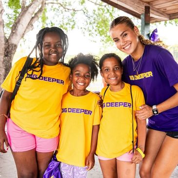Four smiling girls wearing 'DIG DEEPER' shirts, standing under a pavilion.