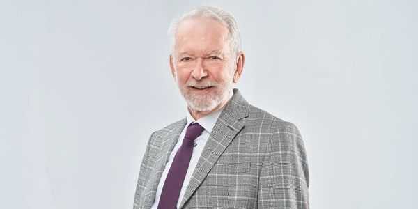 Elderly man in a gray checkered suit and purple tie smiling.