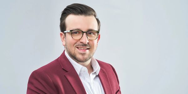 Smiling man in glasses and maroon blazer against a light background.