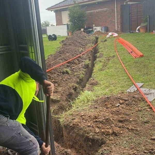Worker digging a trench with orange pipes nearby.