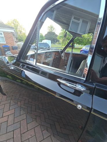 Close-up of a shiny black vintage car door with reflections of houses and trees.