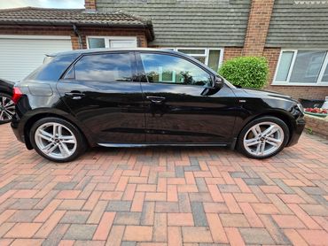 Black Audi hatchback parked on patterned brick driveway beside suburban house.