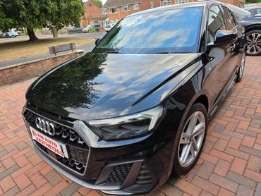Shiny black Audi parked on a brick driveway reflecting trees and sky.