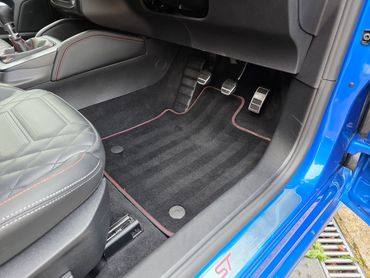 Interior footwell of a blue car with manual pedals and quilted leather seat.