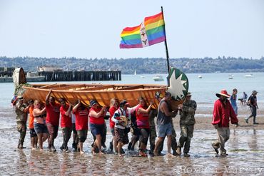 A canoe with a Pride flag is brought onshore by a group of people during Canoe Journey.