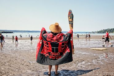 A woman holds a paddle and shows off her Coast Salish regalia onshore during Canoe Journey.